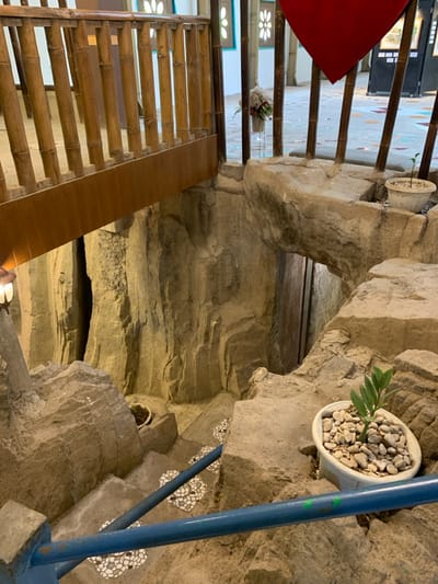The image shows a stone-like, man-made structure resembling a cave, viewed from above. The structure has a small opening that leads down to a set of stairs made of concrete, tiled with white, round pieces. There are two potted plants situated on the upper edges of the structure, one with a green plant and the other with a bushy green plant. To the left of the opening, there is a railing made of bamboo, positioned on a wooden walkway above the structure. The image is likely taken in a park or garden, perhaps a theme park. The coordinates -7.606 latitude and 110.18 longitude correspond to a location within Kecamatan Borobudur in Indonesia.