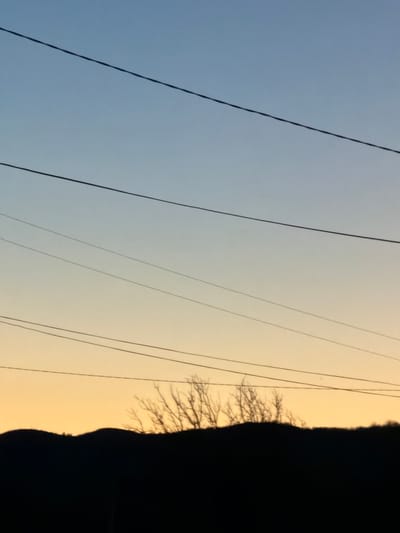 The image shows a view towards the sky in Zlatna Panega, Bulgaria. Power lines stretch across the sky. A dark silhouette of trees and a hill can be seen on the right side. The sky is a gradient of blue on the left, fading to yellow near the right.