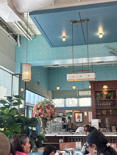 This image depicts the interior of a restaurant or cafe, situated in Highland Park, United States.
The upper sections of the walls feature light blue-green wallpaper with a subtle, repeating pattern of small, lighter-toned graphic motifs. A section of exposed red brick wall is visible behind a service counter. The ceiling integrates both exposed white industrial elements, including ductwork, and a distinct, rectangular light-blue panel containing recessed lighting.
Multiple lighting fixtures are present: a cylindrical pendant light with ribbed sides hangs from a metal chain; a prominent rectangular pendant light with a white diffuser is suspended by two vertical rods from the blue ceiling panel; and several smaller spherical globe pendant lights are distributed throughout the space.
In the midground, a dark wooden shelving unit functions as a bar back, displaying glassware, small floral arrangements, and various decorative objects. Beneath this unit, a light-colored service counter is dominated by a large, ornate vase brimming with a voluminous arrangement of pink, peach, and white flowers. Adjacent to the floral display, commercial kitchen equipment, specifically a coffee machine and a stainless steel ice/drink dispenser, are visible. The wall above the counter exhibits framed artworks, including a landscape and a modern piece with an orange conical figure, alongside a small oval mirror.
The foreground shows three individuals seated at a table. On the left, a woman with long brown hair is visible in profile, looking towards the right. On the right, a woman with dark hair, wearing glasses and a large gold heart-shaped earring, is partially obscured. The top of a child's head is partially visible between these two women. On the table are menus, glassware, and a white paper bag labeled "Pastry Bakery." In the background, behind the service counter, a staff member in a white uniform is partially visible, leaning over the counter.