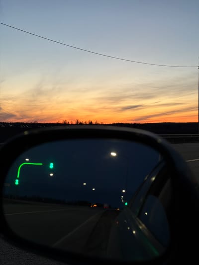 The image presents a dual scene: a sunset vista contrasted with the reflection in a car's side mirror. The sunset displays a gradient of colors, from light blues/grays to oranges/reds, likely indicating the end of the day. A tree line obscures the horizon beneath the sunset. A power line traverses the sky.
The car's side mirror reflects a dark road with streetlights and possibly traffic signals, including a set of two green lights and an artificial turn signal. The scene within the mirror has a distinctly nighttime or very late evening ambiance.
There is nothing in the image to confirm this location in the city of Kuregovo, Russia.