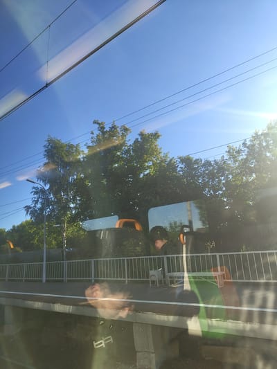 This image depicts a view from a train window, likely at a train station in Konobeevo, Russia, on a clear, sunny day. The sky is a vibrant blue, with several overhead wires visible. Lush green trees line the background, with some appearing to be birch trees due to their slender trunks and lighter foliage.
In the foreground, the train platform is visible, bordered by a white metal fence. Several benches are present on the platform, some with orange accents. Reflections on the train window obscure some details, but the silhouette of a person wearing a dark cap is visible, along with the top of a head with dark hair. Other reflections suggest the presence of more people and possibly parts of the train's interior. The platform surface appears to be made of concrete and gravel. The overall scene suggests a typical day at a suburban train station.