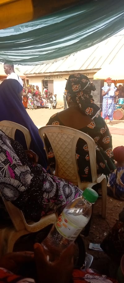 The image depicts an outdoor gathering in Kasuwan Magani, Nigeria, observed from beneath a temporary shelter constructed with green fabric. In the immediate foreground, a dark-skinned hand holds a clear plastic bottle of 7 Up soda with a green cap. The person holding the bottle is seated, partially visible, wearing a dark fabric adorned with white and magenta floral patterns. To their right, another individual is seated with their back to the viewer, wearing a dark green fabric with orange crown-like patterns and a matching headwrap. To the left, a third seated individual is partially visible from the side, wearing a dark blue head covering over a dark garment. All visible seated individuals occupy white plastic chairs.
In the midground, a group of approximately 10-15 individuals, including adults and children, are gathered in an open, unpaved dirt area. Some are seated on the ground or low structures, while others stand, dressed in various patterned traditional attires. To the far right of this group, a woman in a light blue patterned dress and a red headwrap stands near a black speaker and a large traditional drum, holding papers, indicating she may be speaking or presenting.
The background features a single-story building with a light-colored exterior and a corrugated metal roof. The word "MODERN" is distinctly painted on the building's wall. The overall scene is brightly lit by natural light.