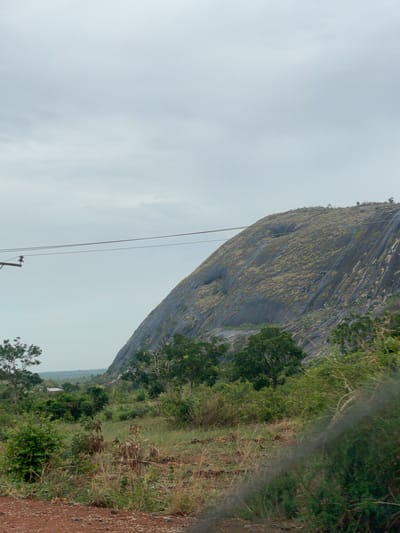 The image shows a large rock formation with a grey, weathered surface, possibly a mountain, located in Kujama, Nigeria. Patches of vegetation cover parts of the rock face. To the right of the rock formation, there is a landscape with trees and grass. On the left, a cloudy sky is visible. A utility pole and wires are positioned in the foreground. The image appears to be taken from a low angle, with the camera tilted. The immediate foreground shows a dirt or gravel surface.