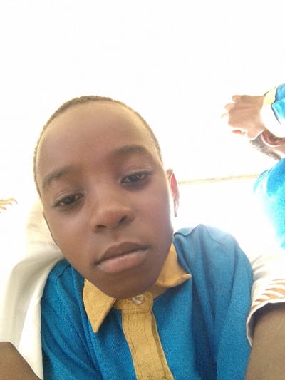The image features a close-up, slightly low-angle portrait of a young boy. He has dark skin and short, dark hair. His gaze is directed forward, meeting the viewer's eyes. His expression is neutral, with his lips gently closed.
He is wearing a blue shirt with a mustard yellow collar and a matching yellow placket (the strip of fabric where buttons are attached). One button at the top of the placket is visible and appears undone. A white or light-colored fabric, possibly a cloak or another piece of clothing, is draped over his left shoulder and arm, extending out of the frame on the left.
To the right side and slightly behind the main subject, another person is partially visible. This person is wearing a blue garment, similar in hue to the boy's shirt, and their arm and hand are visible. The hand appears to be raised near their face or head, and part of a sleeve or arm with a pattern (possibly white with golden or light brown stripes) can be seen.
The background is largely overexposed and appears as a bright, uniform white, suggesting strong, possibly overhead, lighting. This bright background makes it difficult to ascertain the specific setting, though it seems to be indoors. The overall impression is a casual, close-up photograph or a selfie. No text is visible in the image.