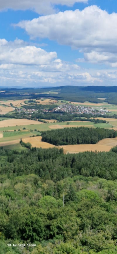 The image is an aerial view of a rural landscape with a town nestled in the valley. The foreground is dominated by dense green foliage, likely trees. In the mid-ground, there is a village with red-tiled roofs and white buildings, surrounded by fields of varying colors, suggesting agriculture. The fields are interspersed with patches of dark green forest. The background features rolling hills covered in dark green forests, with a hazy sky above, filled with white, puffy clouds. The timestamp "19. Juli 2026 14:44" is visible in the lower left corner. The scene appears to be a pastoral view of Neustadt am Kulm, Germany.