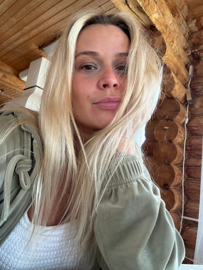 A medium shot of a blonde-haired woman tilted on her side, looking directly at the camera. She is wearing a light green jacket over a white shirt. The background features wooden logs arranged to form a wall or ceiling. Based on the context, this image was taken in Нечкино, Russia.