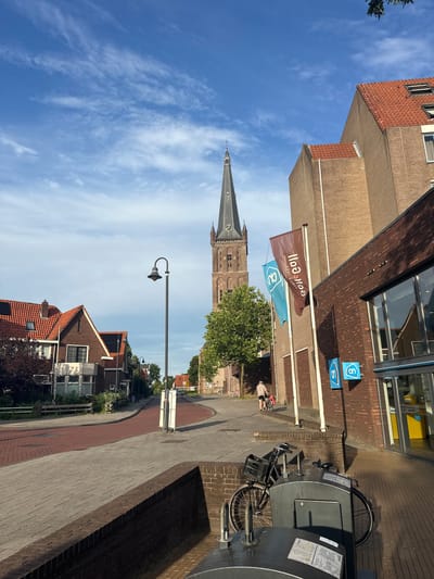 The image depicts a street scene in Steenwijk, Netherlands, on a sunny day with scattered clouds. In the foreground, a brick wall runs along a paved walkway. To the left, a bicycle with a basket is parked near a brick structure. Further down the street, a person is seen pushing a bicycle.
The street curves to the left, lined with red brick houses with tiled roofs. A lamppost stands on the right side of the road. In the background, a tall, dark church tower with a spire and clock dominates the skyline. Adjacent to the church is a modern building with a brick facade, featuring large windows and signage for "Albert Heijn," a Dutch supermarket chain. Two flags, one brown and one blue, with "AH" logos are displayed in front of the building. The sky is a clear blue with wispy white clouds. The overall atmosphere is calm and picturesque, characteristic of a European town.