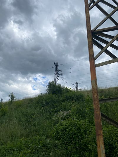 In the city of Lahošť, Czechia, an outdoor scene unfolds under a cloudy sky, dominated by a stark contrast between industrial structures and lush, overgrown vegetation. In the foreground, a large, rusty metal pylon, part of a power line support structure, stands prominently on the right side of the frame. Its weathered beams create a geometric lattice against the overcast sky.
Beyond the pylon, a grassy embankment rises, covered in dense green foliage. Tall grasses and various plants create a wild, untamed appearance. Several electrical pylons, some appearing as slender metal towers with insulators and overhead wires, are visible further up the embankment. These pylons extend into the distance, disappearing behind the curve of the hill. In the background, the sky is filled with heavy, dark clouds, with only small patches of lighter sky and scattered blue visible. The overall impression is one of a somewhat industrial landscape encroached upon by nature. There are no people or animals present in the scene.