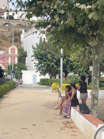 The image shows a street scene in Sant Pol De Mar, Spain, on a paved path lined with a manicured hedge. Several people are present. In the foreground, three women are seated on a low, concrete bench with a red trim. One woman is wearing a black tank top and a patterned skirt, another a black swimsuit, and a third a pink top. Behind them, a fourth woman stands holding a drink. Further down the path, two people, possibly a couple, are walking away from the camera, one carrying a bag. In the background, buildings with white walls and red accents are visible, as well as a stone embankment with arches. The environment is outdoors with trees and foliage, suggesting a sunny day.