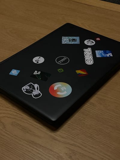 A black laptop with several stickers is placed on a light brown wooden surface. The laptop is slightly ajar, with the screen not visible. The stickers on the laptop include a "NO STRESS" sticker with a water-like background, a round sticker with a floral design, a recycling symbol, a Dell logo, a sticker with the word "tumz" in white outlined text, a sticker depicting a sky with clouds and the text "SAVE THE PLANET", a small green sticker with a circular design, a square sticker with abstract colorful brush strokes, a circular sticker with a white graphic on a gradient background that transitions from orange to blue, and a black and white sticker of a person wearing a hat and holding something. The scene appears to be an indoor setting, possibly a desk or table, in Lomas de Carrasco, Uruguay, given the instruction, but there is no visual information to confirm this specific location or context within the image itself.