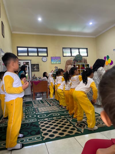 The image captures a scene inside a classroom located in Wonosobo, Indonesia. A group of young children, dressed in yellow and white uniforms, are seated on a patterned green rug. The children are arranged in a line. Behind them, there are shelves, windows, and wall decorations, including a clock and framed pictures. Teachers wearing hijabs are supervising the children. The overall environment suggests a learning space in a school or kindergarten.