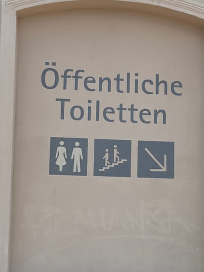 The image shows a sign with German text and symbols indicating public toilets. The text reads "Öffentliche Toiletten," which translates to "Public Toilets." Below the text are three square icons. The first icon depicts a female and male figure, representing gender-neutral or unisex toilets. The second icon shows two figures, one larger and one smaller, ascending a staircase, indicating accessible toilets or a toilet located via stairs. The third icon is a downward-pointing arrow, suggesting the toilets are located below the current level. The sign is mounted on a beige-colored wall. The overall context suggests this is a public facility. Without further information, the exact location in Wismar, Germany cannot be determined.