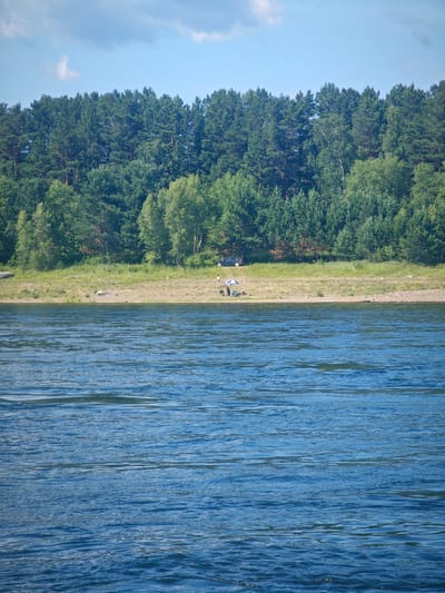 In Abalakovo, Russia, a serene landscape unfolds featuring a wide, calm river in the foreground. The water's surface is a textured tapestry of blues and greens, with subtle ripples and glints of light. Beyond the river, a sandy shore leads to a verdant embankment, carpeted with dry grasses and dotted with scattered rocks. Further back, a dense forest of pine and deciduous trees forms a natural barrier, their foliage a vibrant mix of dark greens and lighter shades. The sky above is a pale blue, streaked with thin, wispy clouds.
On the embankment, a small group of people are enjoying a leisurely day. Two individuals, one appearing to be an adult and the other a child, are near a beach umbrella, suggesting a moment of relaxation or picnic. A dark-colored car is parked discreetly among the trees, hinting at their mode of arrival. The overall scene evokes a sense of tranquility and a connection with nature, common in rural settings like Abalakovo.