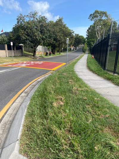 A daytime photograph captures a view along a suburban street in Riverhills, Australia. The asphalt road, marked with double yellow lines, features a prominent red-painted section with white capital letters clearly stating "NO STANDING." A concrete curb separates the road from a grassy verge, which exhibits some patches of drier, sparser grass amidst the green. Parallel to the road, a concrete sidewalk is visible, bordered on the left by a tall, dark metal fence.

Large, leafy trees line the street, casting some shadows, suggesting a sunny day with a clear blue sky dotted with white clouds. In the mid-ground and distance, other houses, fences, and various unreadable road signs can be seen. Several green and yellow wheelie bins (trash cans) are placed along the street, and a small orange traffic cone is visible near the fence further down. Street lamps or light poles are also present. No people or active events are depicted, presenting a static, quiet residential scene.