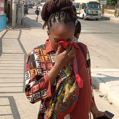 A woman with dreadlocks pulled into a bun stands on a sidewalk. She wears a colorful, patterned shirt and rust-colored pants. She holds a red cloth to her face. A colorful scarf drapes around her neck. A phone is visible in her hand. A busy street with cars and pedestrians is in the background. The scene is brightly lit with sunlight.