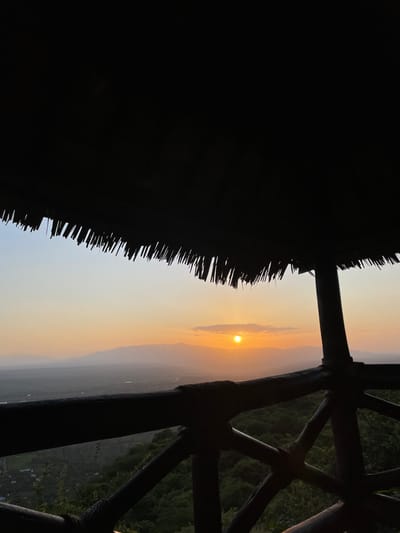 The image captures a serene sunrise or sunset viewed from an elevated, sheltered vantage point in Mto Wa Mbu, Tanzania. The scene is dominated by warm, hazy light characteristic of dawn or dusk.
In the foreground, the top of the frame features the dark, silhouetted edge of a thatched roof, its individual strands clearly visible. On the right side, a rustic wooden railing or part of an open-air structure, composed of dark, crisscrossing beams, is also visible in silhouette, framing the expansive view.
The sun, a brilliant golden orb, is positioned on the left side of the image, just above the hazy horizon. The sky exhibits a stunning gradient, transitioning from a soft light blue and grey in the lower left to vibrant oranges and yellows around the sun, fading into paler tones higher up. Distant mountains or hills form a layered silhouette along the horizon line, partially obscured by atmospheric haze. Below these distant landforms, a vast valley or flat landscape extends into the distance, appearing dark and muted under the backlighting.
The image conveys a sense of tranquility and awe, highlighting the natural beauty of the Tanzanian landscape at a pivotal time of day. No visible text is present in the media file.