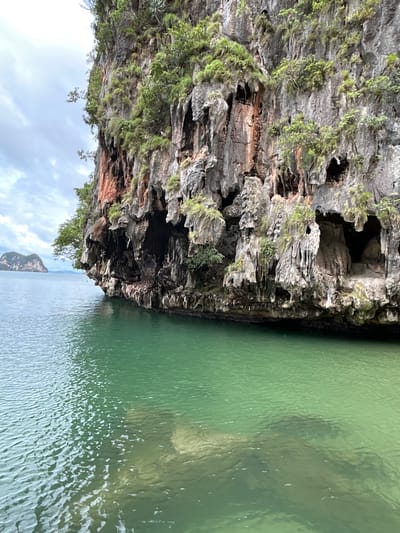 The image depicts a dramatic natural landscape featuring a towering, rugged limestone cliff face overgrown with lush green vegetation. The cliff has numerous natural caves and overhangs, with stalactite-like formations dripping from the edges. The rock exhibits varied colors, including shades of gray, brown, and striking patches of reddish-orange. Below the cliff, the clear, emerald-green water of the sea gently laps against its base. In the distance, to the left, a smaller, tree-covered island can be seen rising from the water under a partly cloudy sky. The perspective suggests the photo was taken from a boat or a lower vantage point near the water's surface. The scene evokes a sense of natural beauty and geological wonder, characteristic of coastal areas in Phang Nga Bay, Thailand, such as those found in Tambon Krasom. No people or man-made structures are visible.