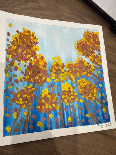 A painting on white paper depicts a forest with trees with sparse foliage in warm autumn colors of yellow and brown, set against a bright blue sky with vertical brushstrokes. Scattered leaves in similar fall colors are depicted floating in the air. The style is Impressionistic, with dabs of paint used to create the impression of leaves and branches. The bottom right corner of the painting has a handwritten date "15.11.23". The painting is on a wooden surface, possibly a table. There are no people or animals visible in the painting.