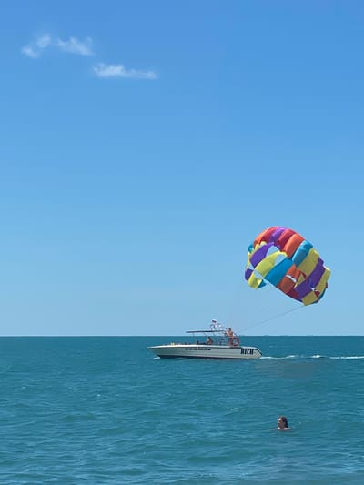 The image captures a sunny day at sea, with a clear blue sky and calm turquoise waters. The main subjects are a parasailing setup and a boat. A brightly colored parasail, a vibrant mix of yellow, purple, blue, orange, and red stripes, is visible in the sky, tethered to a boat. The boat, named "RICH", is white with dark trim and appears to be a speed boat designed for watersports. Several people are visible on the boat, including a few passengers and at least one crew member operating the equipment.

In the water, a person is swimming, their head and shoulders visible above the surface. The water has gentle ripples, indicating a tranquil environment. A few wispy clouds are present in the sky, adding a touch of texture to the otherwise clear expanse. The overall scene suggests a recreational activity, likely parasailing and swimming, taking place in a coastal or open sea location, consistent with Adlerskiy, Russia. The bright sunlight suggests it is daytime, possibly late morning or afternoon, with good weather. Visible text on the boat includes "RICH" and what appears to be a registration number or identifier, "BE 01 06 ROS 23-". The atmosphere conveyed is one of leisure and outdoor fun.