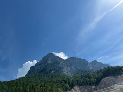 A wide, low-angle shot captures a majestic mountain range under a clear, bright blue sky. The mountain's face is steep and rocky, with visible striations and shadows that highlight its rugged texture. Lush green forests carpet the lower slopes of the mountains, their dense foliage contrasting with the grey rock. A few white, fluffy clouds are scattered across the sky and near the mountain peaks. In the upper right of the image, a thin, wispy contrail stretches across the sky, suggesting air traffic. The bright sunlight indicates it is daytime, likely mid-morning or afternoon. The scene evokes a sense of natural grandeur and tranquility. The location is Schneizlreuth, Germany. No text is visible in the image.