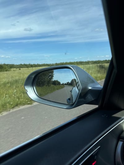 The image shows a view from inside a car, looking out the side window and into the side-view mirror. The scene is dominated by a bright blue sky with scattered white clouds, suggesting it is daytime and the weather is fair. To the right of the mirror, a section of the car's interior is visible, including part of the dashboard and door panel, rendered in dark grey. The main focus is the side-view mirror, which reflects a portion of the road and the surrounding landscape. Through the mirror, trees and green fields can be seen lining the road, indicating a rural or semi-rural setting. The car appears to be in motion, as suggested by the blurred background and the perspective of the reflection in the mirror. The overall impression is one of travel through a scenic countryside under a pleasant sky. The image appears to be taken in Dovhe, Ukraine. No people are visible in the image. No text is visible in the image.