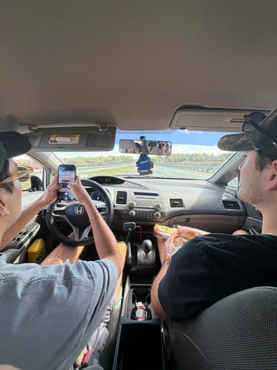 Two adult males are depicted in the front seats of a moving vehicle, a Honda Civic, on a multi-lane highway near Gainesville, United States. The interior perspective is from the back seat, looking forward.
The driver, positioned on the left, wears a dark baseball cap, light-colored short-sleeved shirt, and shorts. He holds a black smartphone horizontally with both hands, appearing to photograph or record the road ahead. A gold bracelet is visible on his right wrist. Sunglasses are perched on his cap.
The front passenger, on the right, wears a camouflage-patterned baseball cap and a dark short-sleeved shirt. He is holding a clear plastic bag containing multiple bread rolls in his lap.
The vehicle's dashboard, featuring a Honda steering wheel, is visible. The speedometer reads "67". A blue "Little Tree" air freshener hangs from the rearview mirror, which reflects the front occupants. The center console includes a manual gear shifter, a blue electronic device plugged into a port, and a cup holder containing a small red and white box. A yellow container is present in the driver's door pocket.
Outside the vehicle, a multi-lane asphalt highway is visible under a clear blue sky with some white clouds. Green vegetation lines both sides of the road, and a guardrail is visible on the left.