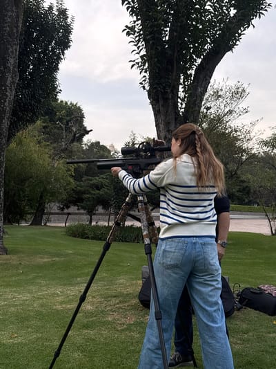 A woman stands in a grassy field, aiming a rifle mounted on a tripod. She wears a striped sweater and jeans. An older man is behind her. The backdrop includes lush trees and foliage. The scene appears to be outdoors on a cloudy day.