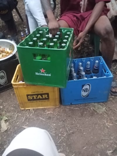 Three crates of bottled beverages, stacked unevenly, sit on bare ground. The top crate, green and bearing a "Heineken" logo, is full. Below it, a yellow crate displays "STAR," while a blue crate labeled "NB" sits to the side. Partially visible people flank the crates, one wearing a maroon outfit and sitting on a green stool. There's also an individual wearing white pants with tattooed arms. A metal bowl filled with food is to the left, on a black crate.