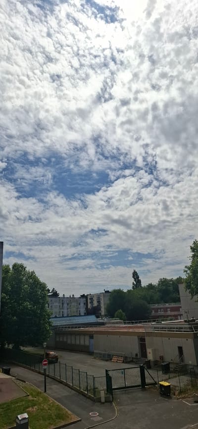 The image shows a cloudy sky over an urban environment in Le Havre, France. The sky is filled with white, fluffy clouds interspersed with patches of blue. The clouds are dense and appear to be at a medium altitude, suggesting partly cloudy or overcast conditions.
Below the sky, there are buildings and trees. The buildings are of varying heights and architectural styles, with some appearing to be apartment blocks. One building has a red brick facade on the upper floors. There is also a building with a modern, glass-enclosed lower level. Trees are scattered throughout the scene, with some appearing to be mature and leafy, providing greenery against the urban structures.
In the foreground, there is a paved area, possibly a street or courtyard, with a black metal fence bordering it. There is a yellow dumpster and a red and white "no entry" sign visible near the fence. Further to the right, there is a patch of grass. The scene suggests an outdoor setting in a residential or mixed-use area.
The lighting indicates it is daytime, likely late morning or afternoon, given the brightness of the clouds and the shadows cast. The overall mood is somewhat neutral, reflecting a typical day in a city with changeable weather. No people are clearly visible in the image, but there is a small, dark object that could be a car parked on the side of the paved area. There is no visible text in the image other than on the "no entry" sign.