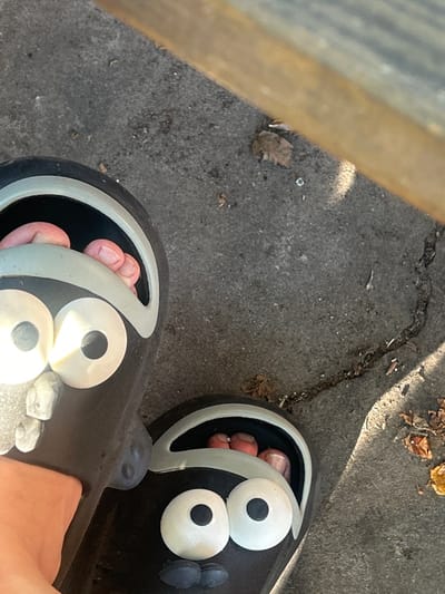 The image, captured in Dibrova, Ukraine, presents a top-down view of a person's bare feet wearing two black slip-on sandals. Each sandal features a stylized face design on the strap, consisting of two large white circular eyes with black pupils, a small gray rounded nose, and a small black mouth. The toes of both feet are visible within the sandals. The sandals rest on a coarse, cracked paved surface, consistent with concrete or asphalt, which is strewn with scattered dry leaves and small debris. A light-colored wooden structure is partially visible along the upper right edge of the frame, and direct sunlight casts shadows on the ground surface.