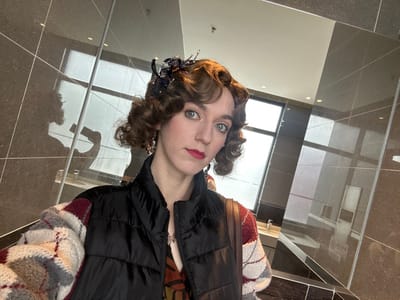 A female individual is depicted in a selfie taken within a modern public restroom in Huangdao District, China. The subject has light skin, blue-green eyes, and dark red lipstick on her lips. Her brown hair is styled in prominent curls, adorned with a dark, beaded accessory on the right side of her head. She wears reddish earrings, a black puffer vest over an argyle-patterned sweater in red, white, and gray, and an underlying patterned top. A brown bag strap is visible over her right shoulder. The background consists of large, dark brown or charcoal-colored glossy wall tiles, which reflect the individual holding a smartphone to capture the image. A large window to the left reveals a bright exterior view, likely an overcast sky or an outdoor structure. Reflections on the tiled surfaces also show a white sink with a chrome faucet on a dark countertop, and a light-colored ceiling with recessed lighting.