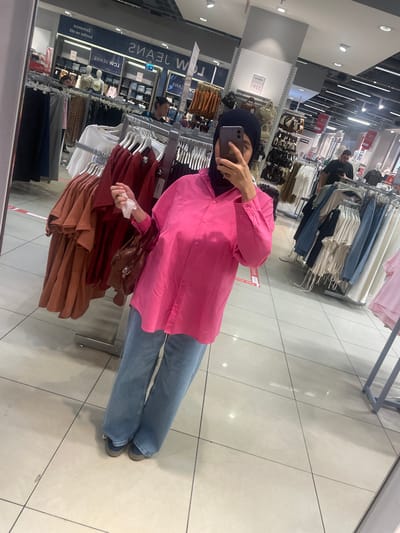 A person wearing a navy blue hijab and a bright pink oversized button-down shirt is taking a mirror selfie in a clothing store. They are also wearing light-wash blue jeans and dark-colored shoes. In their left hand, they are holding a white tissue or small paper. A brown crossbody bag is slung across their body. The background shows racks of clothes for sale, including jeans, t-shirts, and dresses. The store appears to be well-lit, with fluorescent lighting on the ceiling. Reflections in the mirror show more clothing racks and other shoppers in the distance. The floor is tiled with a light-colored ceramic tile. The overall impression is of a casual shopping trip. Visible text includes "LCW JEANS" on a sign in the reflection.