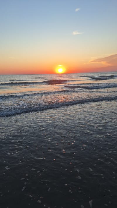 The image depicts a serene sunset over the ocean at the beach in Berck, France. The sun, a bright orange orb, is low on the horizon, casting a warm glow across the sky and water. Gentle waves roll towards the shore, their crests illuminated by the fading sunlight. The foreground is dominated by the dark, glistening surface of the water, dotted with small white bubbles. The sky transitions from a clear, pale blue at the top to a gradient of soft orange and pink hues near the horizon. A few wispy clouds are visible in the upper part of the sky. There are no people or man-made objects in the frame, creating a natural and tranquil scene.