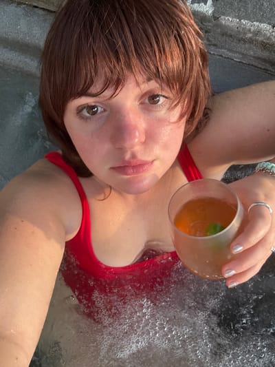 A woman with brown hair and eyes poses in a hot tub while holding a glass of amber-colored liquid. She wears a red bathing suit and turquoise and silver jewelry. Her hand has several silver rings and glittery nails. Bubbles surround her chest in the water. She looks up at the camera with a relaxed expression.