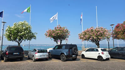 The image captures a sunny day in Sirmione, Italy, with the shimmering waters of Lake Garda providing a backdrop. Several cars are parked on a cobblestone street lined with trees. A black SUV is parked on the left, followed by a silver Citroen hatchback. In the center, a light blue Land Rover Defender is parked, featuring a spare tire mounted on the rear. To the right, a white Smart car is parked, and a dark grey SUV is partially visible on the far right.

Several flags are hoisted on tall poles along the waterfront. From left to right, the flags visible are the European Union flag, the Italian flag, and a flag with red and white stripes on a blue background, possibly a regional or nautical flag. Two trees with white blossoms and two trees with pink blossoms are prominently featured, adding vibrant color to the scene.

In the background, the blue waters of Lake Garda are calm, with a large white cruise ship visible in the distance. The shoreline is dotted with buildings and hills, characteristic of the Sirmione landscape. The sky is a clear, bright blue with a single small bird flying high above. Parking signs are visible near the trees, indicating parking regulations.