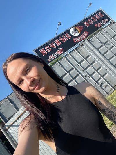 A selfie of a woman with dark hair and tattoos on her arms and neck is centered in the frame, with a bright blue sky visible in the background. Behind the woman is a large sign with the words "HOYLE ROCK RODEO" written in a stylized font, along with a graphic of a wolf and flames. The sign is attached to a metallic structure, possibly a building or a large storage container. The location is identified as Doma 45 Kilometr, Russia.