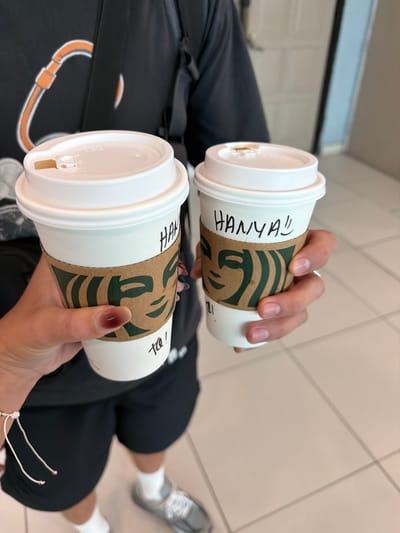 Two Starbucks cups are held in a person's hands. The cups have white lids and sleeves with the Starbucks logo. The name "HANYA!!" is written on one cup, and "Ta !" is written on the other. The person holding the cups is wearing black shorts and a black bag. A pair of silver sneakers are visible in the background. The background appears to be an indoor tiled floor. The location cannot be determined to be in George Town, Malaysia from the image alone.