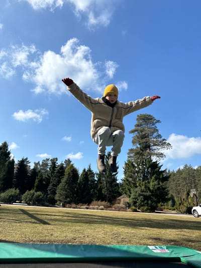 A child in a yellow beanie, beige jacket, gray pants, and black boots is in mid-air against a blue sky with scattered clouds. The child's arms are outstretched and they are wearing red mittens. To the right of the child, there's a grassy field, trees, and the edge of a trampoline. In the background, more trees, a white car, and a basketball hoop are visible. The location is Stapriņi, Latvia.