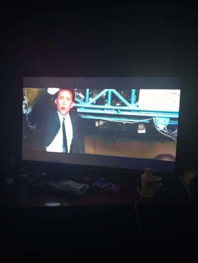The image shows a television screen displaying a scene from a movie or show. A man in a suit and tie is visible on the screen, with his mouth open. Behind him, there is a blue metal structure and some wires. The television is on a stand in a dark room. On the stand in front of the television, there are some objects, including a bottle of liquid, some wires, and a folded piece of cloth. The overall scene is dimly lit, with the television screen being the primary source of light. The location is not specified as Suklieia, Moldova.