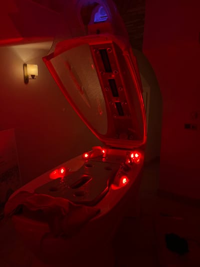 The image depicts a spa treatment room in Kliczków, Poland, illuminated predominantly by red light. The central subject is a white, pod-like spa capsule or hydrotherapy bed with its upper lid fully open, angled upwards. Inside the open lid, three rectangular digital displays or control panels are vertically aligned. The interior of the lower bed section features multiple circular red LED lights that are actively illuminated, casting a strong red hue across the room. Condensation or water droplets are visible on the internal surfaces of both the lid and the bed. A folded, light-colored towel is positioned at the foot end of the lower bed. On the left wall, a wall-mounted sconce with a translucent shade emits a warm, yellowish light. A white poster or sign with dark text is partially visible on the far left wall. The room's walls are light-colored, appearing red under the dominant lighting. An arched doorway or opening is visible on the right side of the frame. A dark object rests on the floor in the lower right corner. No individuals are present in the scene.