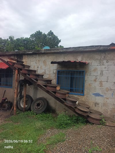 The image shows an outdoor scene in El Chaparral, Venezuela, under an overcast sky. A weathered, light-colored concrete block building with a red corrugated metal awning above a blue-barred window is visible. In front of the building, a makeshift staircase constructed from metal beams and what appear to be repurposed metal wheels or tires ascends diagonally. The staircase is rusted and appears old. Below the staircase, two large vehicle tires are leaning against the building. The ground is covered with grass, gravel, and some low-lying vegetation. The lower left corner of the image has a timestamp and text overlay: "2026/06/19 06:52" and "natha".