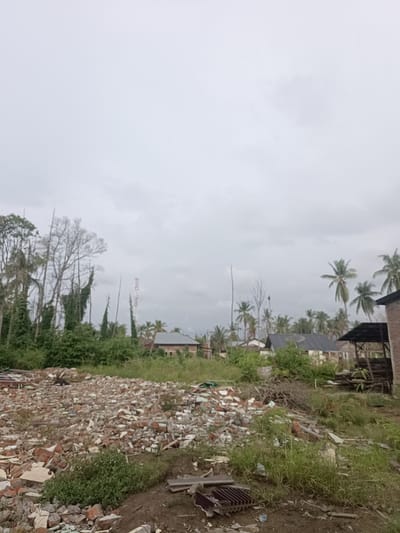The image depicts a cloudy, overcast sky over a scene of destruction and rebuilding in Meunasah Pupu, Indonesia. In the foreground, a large pile of rubble consisting of broken bricks, concrete, and other debris dominates the left side of the frame. Some green vegetation is growing amidst the debris.

In the mid-ground, several structures are visible. To the left, a dense cluster of trees, some with ivy climbing them, stands tall. A red and white telecommunications tower rises behind these trees. Further to the right, several houses with dark roofs are scattered amongst palm trees. One building appears to be under construction or renovation, with exposed brickwork and a covered open-air structure adjacent to it.

The overall impression is one of a community recovering from an event that caused significant damage, with remnants of destruction juxtaposed with signs of ongoing life and development. The environment appears to be a semi-rural or suburban area.