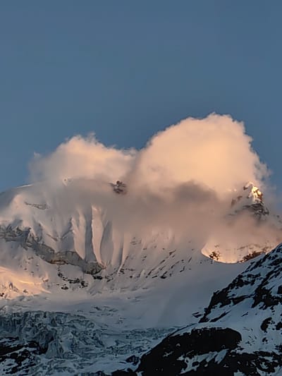 A majestic snow-capped mountain peak is bathed in the warm, golden light of either sunrise or sunset. A thick, fluffy cloud, tinged with the same warm hues, partially obscures the summit, creating a dramatic and ethereal effect. The mountain's rugged slopes are covered in pristine snow, with visible crevasses and the stark contrast of dark, rocky outcrops peeking through. Below the main peak, a glacier is visible, its icy surface textured and reflecting the light. The sky above is a clear, tranquil blue, suggesting fair weather. The overall impression is one of awe-inspiring natural beauty and the raw power of the elements. The time of day is either early morning or late afternoon, given the warm, low-angle sunlight. The location is implied to be a mountainous region, given the snow, glacier, and sheer rock faces.