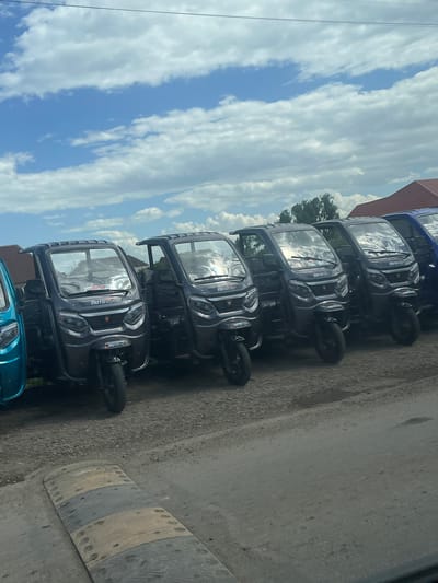 The image depicts a row of approximately five or six three-wheeled vehicles, similar in style to auto-rickshaws, parked along a paved street. The vehicles are mostly dark gray or black with windshields and "Motosafe" logo stickers. The sky behind them is blue with scattered white clouds. The location is Nakhiduri, Georgia. The scene appears to be outside, likely near a dealership or parking area for these vehicles. A small section of a building is visible in the background. A few green bushes are also noticeable.
