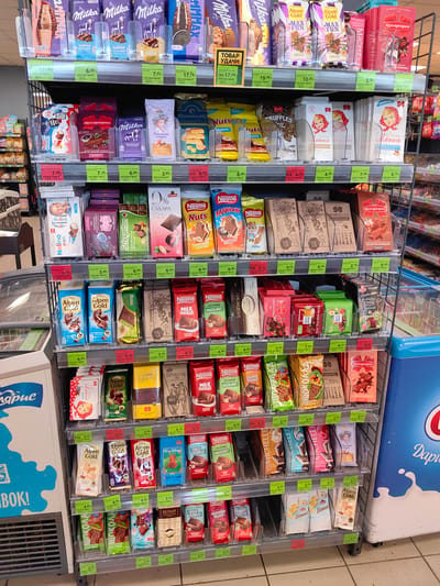 The image shows shelves stocked with a variety of chocolate bars and sweets in a store. The shelves are metal and have price tags below each product. Products visible include Milka, Alpen Gold, Nestle, and Ritter Sport, among others. Some products are individually wrapped, while others are in larger boxes. The lighting appears to be artificial, typical of an indoor retail environment. The overall impression is of a well-organized candy aisle. The location is identified as a store in Myadzyel, Belarus. There are no people visible in the image.