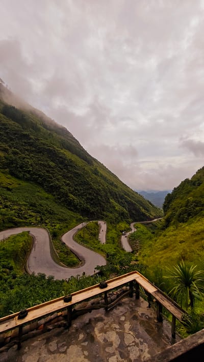 A high-angle shot shows a winding road in Phố Bảng Commune, Vietnam, set against a backdrop of lush, green mountains and a cloudy sky. The road snakes through the valley, with a couple of vehicles seen navigating the turns. In the foreground, a wooden structure, possibly a viewing platform, offers a vantage point to observe the landscape. The platform has a weathered appearance, with evidence of moisture. The overall atmosphere is one of rural tranquility, punctuated by the road's infrastructure.