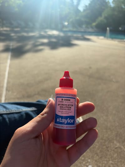 A person's hand holds a Taylor R-0004 pH Indicator Solution (Phenol Red) in an outdoor setting. The solution comes in a red plastic bottle with a red cap, bearing the Taylor logo and product information, including the volume of 60 mL (2 oz). The label also states "KEEP OUT OF REACH OF CHILDREN" and "TEST PURPOSES ONLY - NOT FOR INTERNAL USE." The person is wearing blue jeans. The background consists of an out-of-focus area with hints of greenery, suggesting trees or vegetation, and a lighter-colored surface possibly indicating a sidewalk or pavement. The image suggests the photo was taken outdoors on a sunny day, given the strong sunlight and shadows. The label indicates Taylor Water Technologies LLC at 31 Lovelon Circle. Edison, United States of America.