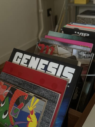 A close-up, canted shot shows a row of records in a wire record rack. The nearest record is a colorful, cartoonish illustration dominated by green, red, yellow, and blue hues. It sits behind a red record cover. Next to this is a record with the word "Genesis" written vertically in large, white letters on a black spine. Other records with varying colors and titles are visible behind it. The context for Saint-Ouen-sur-Seine, France, is absent from the provided information.