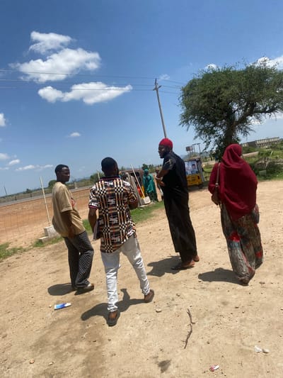 The image depicts a group of people outdoors in what appears to be Yali, Nigeria. The scene is set on a dusty, unpaved ground with a few sparse trees and some distant buildings under construction. The sky is a vibrant blue with scattered white clouds, suggesting a sunny day.
In the foreground, three young men are visible. The man closest to the camera is wearing a light brown t-shirt and patterned trousers, facing left. Next to him, a man in a checkered shirt and white trousers is looking to his left. The third man, further back and slightly out of focus, is dressed in a black thobe and a red cap, with his hands clasped in front of him. Behind him, a woman in a red hijab and a colorful patterned skirt is also present. Another figure in a green garment is partially visible in the background.
The people appear to be standing and possibly engaged in a conversation or waiting. There are some electrical wires and a utility pole visible, as well as a small yellow vehicle in the middle ground with some posters on its side, one featuring a portrait of a person. The presence of these elements, along with the vegetation and architecture, points to a rural or semi-urban setting in Nigeria. The lighting suggests it is daytime, likely mid-morning or afternoon, given the shadows cast on the ground. No specific emotions are discernible from their postures. There is a small blue and white object on the ground in the foreground.