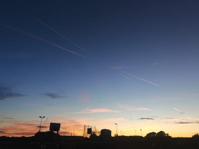 This is a silhouette image of a sunset in Worthing, United Kingdom. The sky is a gradient of deep blue at the top transitioning to orange and pink hues near the horizon. Several contrails are visible in the sky, indicating airplane activity. In the foreground, silhouetted against the sunset, are various structures including lampposts, a tall building with a distinctive angular roof, what appears to be a construction crane, and a large, possibly electronic, sign. Below these structures, the silhouettes of trees and low-lying buildings are visible, along with what looks like recreational equipment, perhaps for a park or sports field. The overall impression is an urban landscape at dusk, with the sky as the dominant element due to the sunset and contrails.
